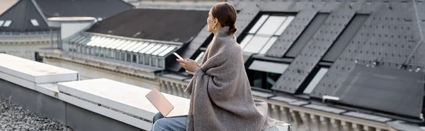 Donna ricoperta di scialle seduta con smartphone e laptop sul tetto, banner — Foto stock