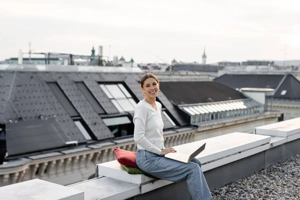 Happy woman looking at camera while sitting on rooftop with laptop — Foto stock