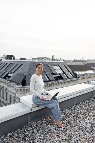 Весела жінка в джинсах сидить на даху з ноутбуком і посміхається на камеру — стокове фото