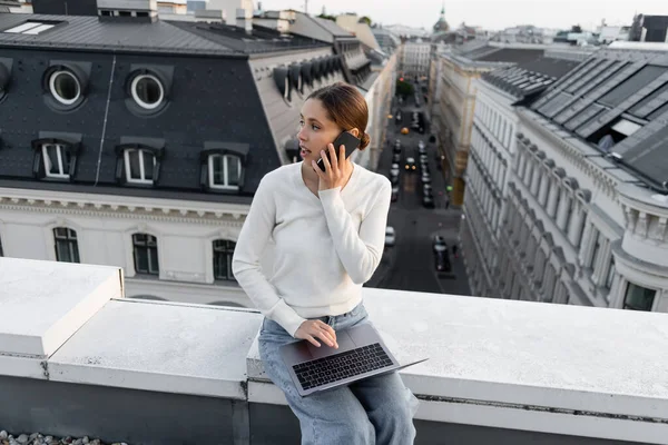 Woman sitting on rooftop with laptop and talking on mobile phone — стоковое фото