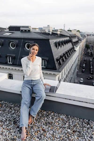 Femme souriante regardant la caméra tout en parlant sur smartphone près d'un ordinateur portable sur le toit — Photo de stock