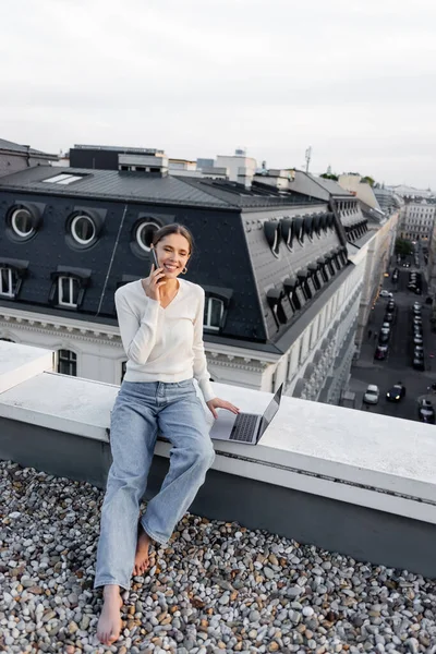 Full length of happy woman talking on smartphone near computer on rooftop - foto de stock