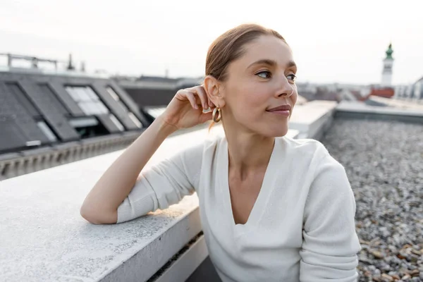 Lächelnde Frau im weißen Pullover, die auf dem Dach eines städtischen Gebäudes wegschaut — Stockfoto