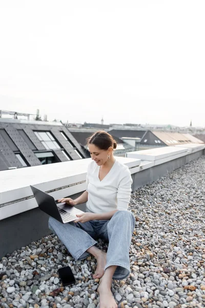 Улыбающаяся босиком женщина, работающая на ноутбуке, сидя на крыше возле мобильного телефона — стоковое фото