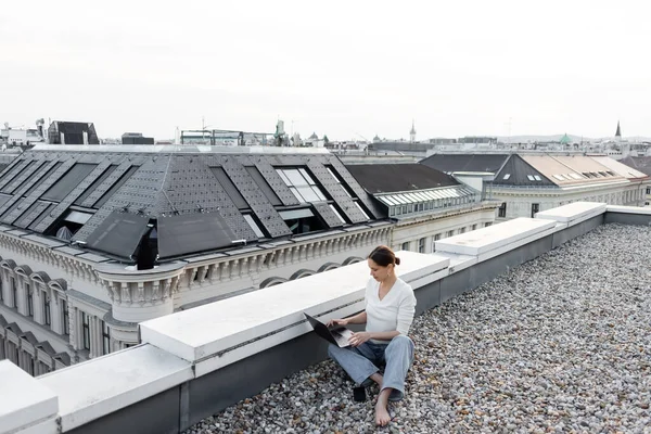Full length of barefoot woman in jeans working with laptop on rooftop of urban building - foto de stock