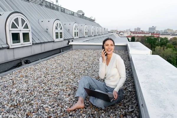 Full length of woman in jeans sitting on rooftop with laptop and talking on cellphone — стоковое фото