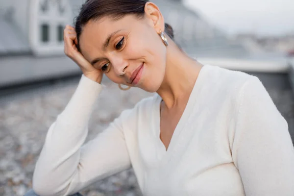 Mujer de ensueño en jersey blanco sosteniendo la mano cerca de la cabeza mientras sonríe al aire libre - foto de stock