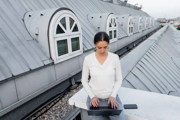 Mujer en jersey blanco escribiendo en el ordenador portátil cerca de teléfono inteligente con pantalla en blanco en la azotea - foto de stock