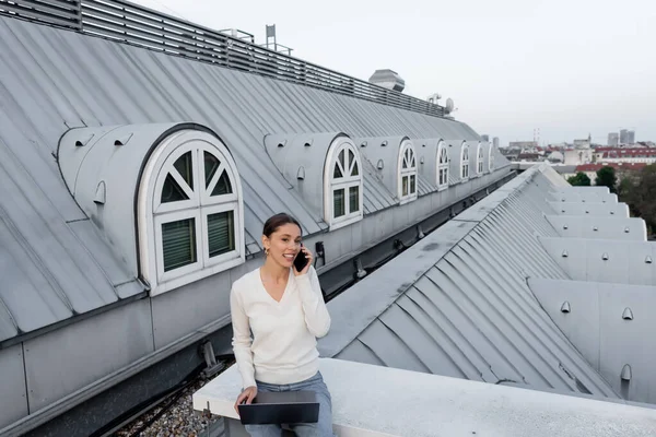 Femme souriante avec ordinateur portable parlant sur le téléphone mobile tout en étant assis sur le toit de l'immeuble de la ville — Photo de stock