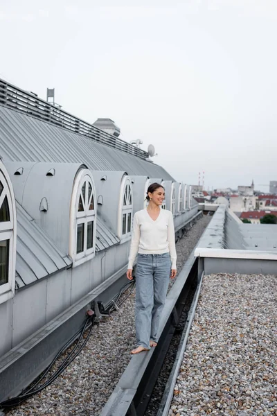 Full length of barefoot woman in jeans walking on rooftop of urban building — стоковое фото