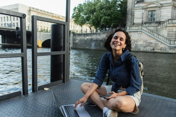 Smiling young freelancer holding smartphone and using laptop near river in berlin — Stock Photo
