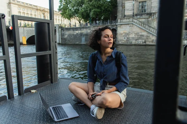 Surprised freelancer holding smartphone while sitting with crossed legs near laptop and river in berlin — Stock Photo
