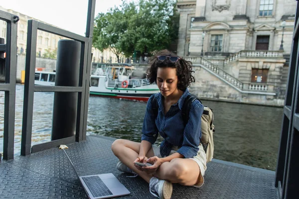 Curly freelancer using smartphone while sitting with crossed legs near laptop and river in berlin — Stock Photo
