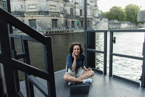 Happy young woman using laptop while sitting with crossed legs near river in berlin — Stock Photo