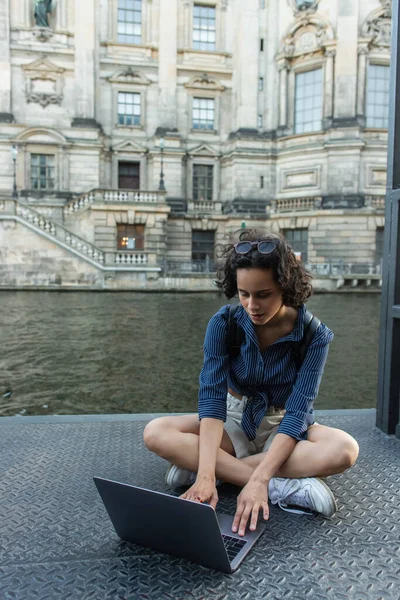 Молодой фрилансер с ноутбуком, сидя со скрещенными ногами возле реки в Берлине — стоковое фото