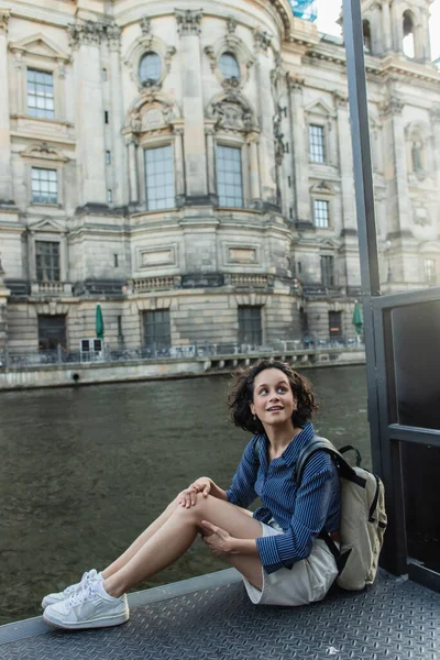 Изумленная и молодая женщина, сидящая возле реки и старинного здания в Берлине — стоковое фото