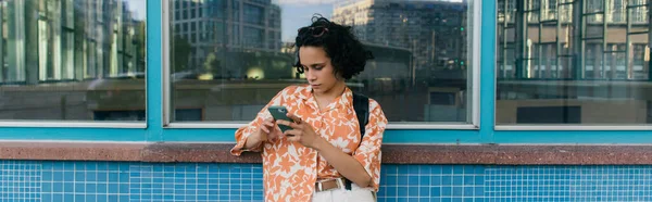 Curly young tourist messaging on smartphone near building in berlin, banner — Stockfoto