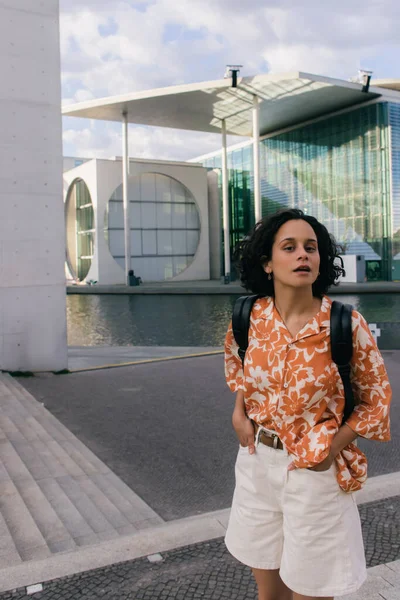 Curly young woman standing with hands in pockets near modern contemporary building in berlin - foto de stock