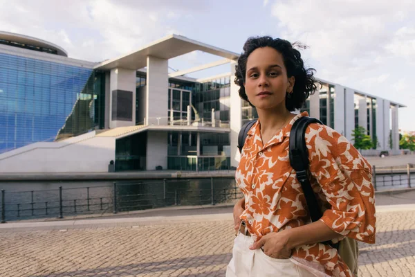 Curly woman with backpack standing with hands in pockets near modern contemporary building in berlin — Stock Photo