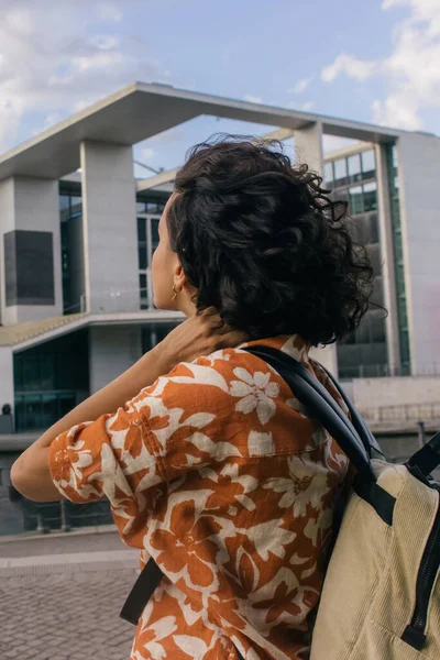 Curly young woman with backpack looking at modern contemporary building in berlin — Stockfoto