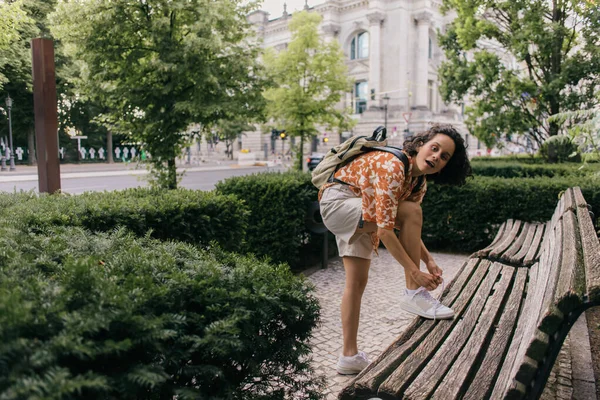 Amazed young woman in summer outfit tying shoelaces in green park in berlin - foto de stock