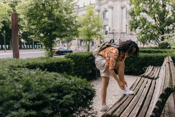 Curly young woman in summer outfit tying shoelaces in green park in berlin — Foto stock