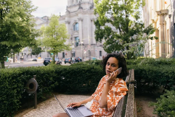 Young freelancer in summer outfit sitting on bench with laptop and talking on smartphone in green park — Photo de stock