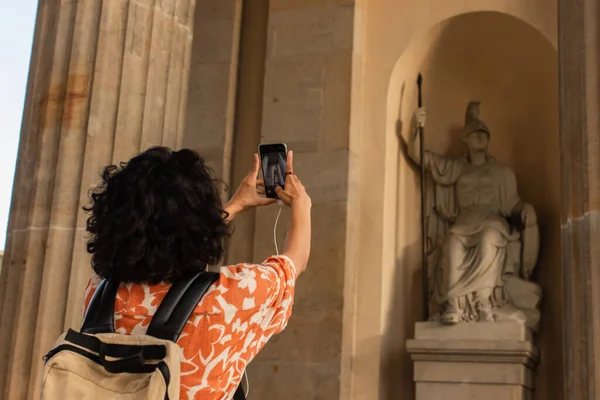 Back view of curly tourist with backpack taking photo of statue on smartphone — Fotografia de Stock