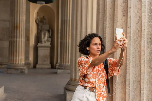 Curly young tourist in wired earphones taking selfie with statue while holding smartphone — Fotografia de Stock