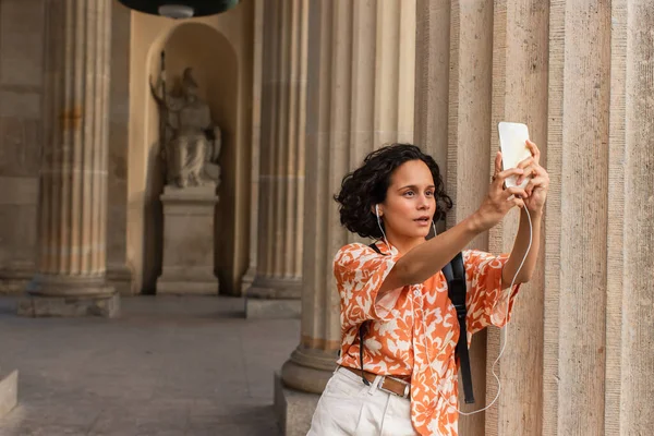 Curly young tourist in wired earphones taking selfie with statue while holding smartphone — Stock Photo