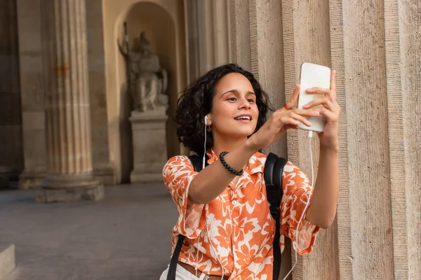 Happy young tourist in wired earphones taking selfie with statue while holding smartphone — Fotografia de Stock