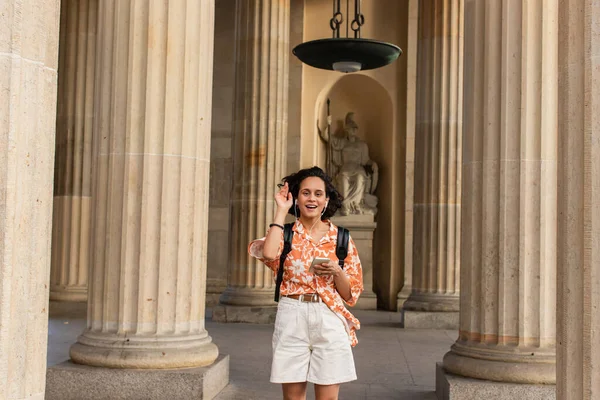 Amazed traveler in wired earphones listening music and holding smartphone near blurred statue in berlin — Fotografia de Stock