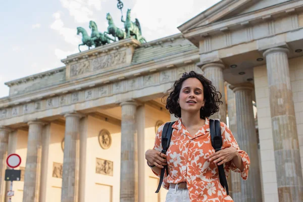 Smiling tourist with backpack standing near brandenburg gate in berlin - foto de stock