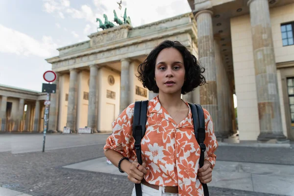 Curly young woman with backpack standing near brandenburg gate in berlin — Stock Photo