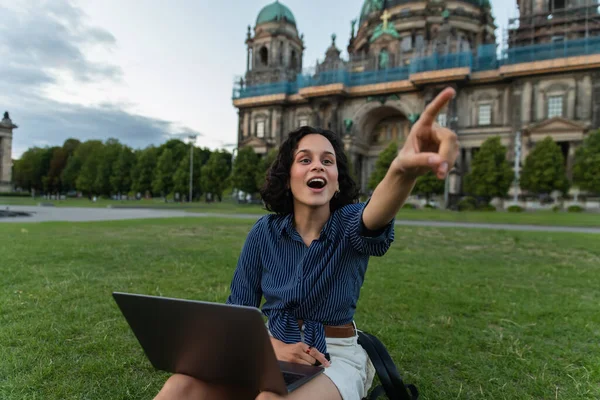 Amazed young woman with laptop pointing with finger and sitting on grass near blurred cathedral in berlin — Foto stock