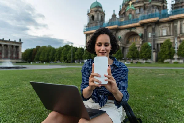 Smiling woman sitting with laptop and taking selfie on smartphone near cathedral in berlin - foto de stock