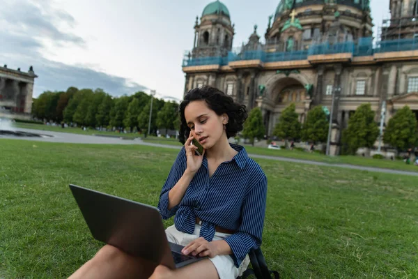 Curly young woman sitting with laptop and talking on smartphone near cathedral in berlin — Foto stock