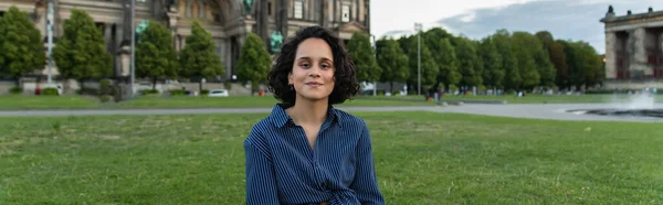 Cheerful young woman sitting on grass near blurred building in berlin, banner - foto de stock