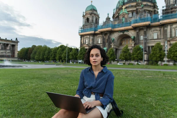 Young freelancer using laptop and sitting on grass near blurred cathedral in berlin during trip — Photo de stock
