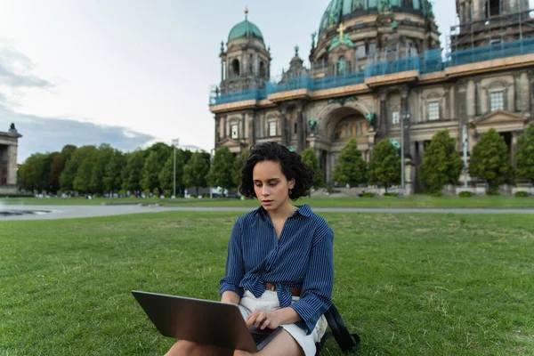 Young woman using laptop and sitting on grass near blurred cathedral in berlin during trip — Foto stock