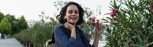 Pleased young woman with backpack touching flower on green bush in berlin, banner — Photo de stock