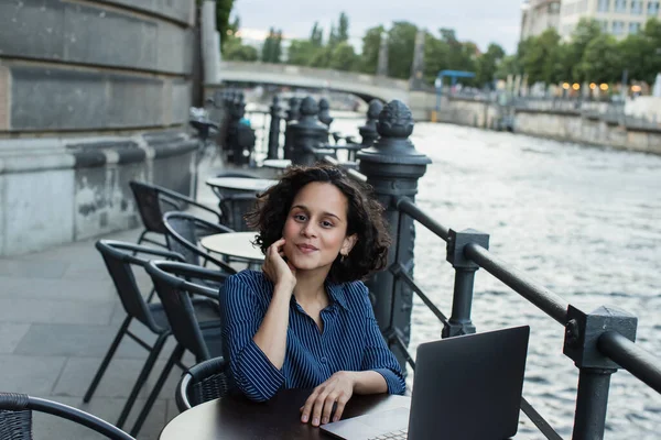 Curly young freelancer using laptop while sitting on summer terrace near river in berlin — Foto stock