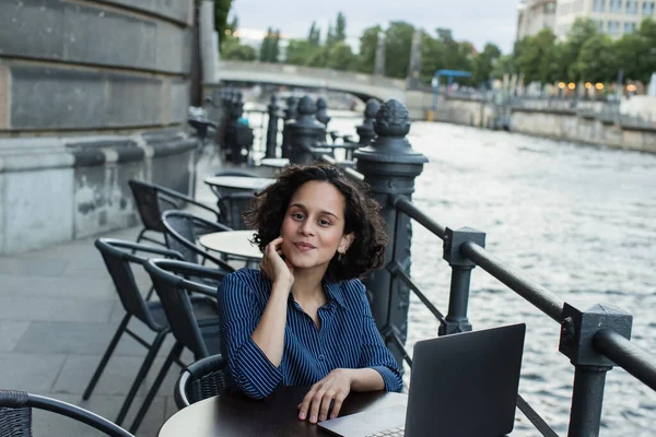 Curly young freelancer using laptop while sitting on summer terrace near river in berlin — Stock Photo