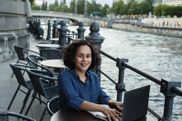 Cheerful young woman using laptop while sitting on summer terrace near river in berlin - foto de stock