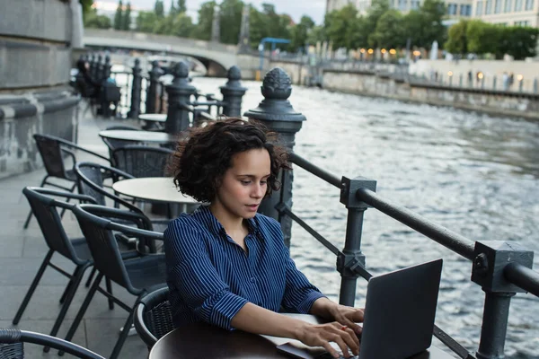 Curly young woman using laptop while sitting on summer terrace near river in berlin — Stock Photo