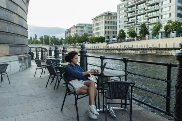 Full length of young freelancer using laptop while sitting on summer terrace near river in berlin - foto de stock