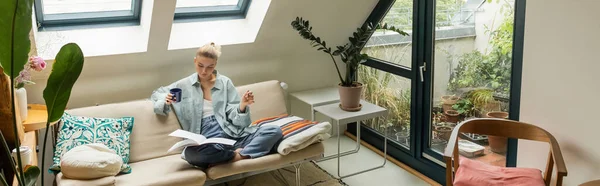 Frau hält Tasse und schaut ein Notizbuch, während sie auf Couch im Wohnzimmer sitzt, Banner — Stockfoto