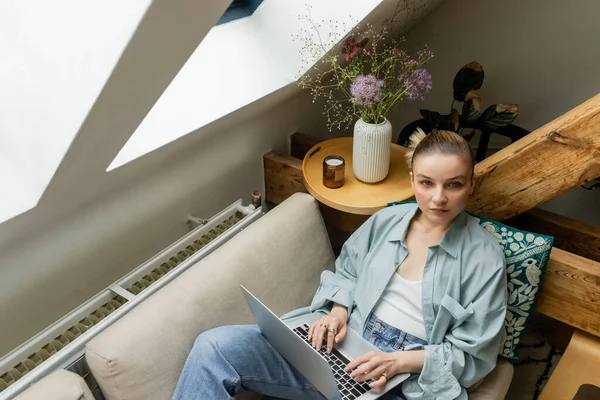 Visão de alto ângulo da mulher olhando para a câmera perto do laptop no sofá na sala de estar — Stock Photo