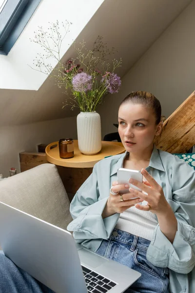 Femme pensive utilisant un ordinateur portable et un smartphone assis sur le canapé à la maison — Photo de stock