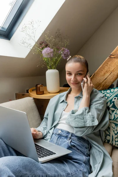 Smiling freelancer talking on smartphone and looking at camera near laptop at home — Stock Photo